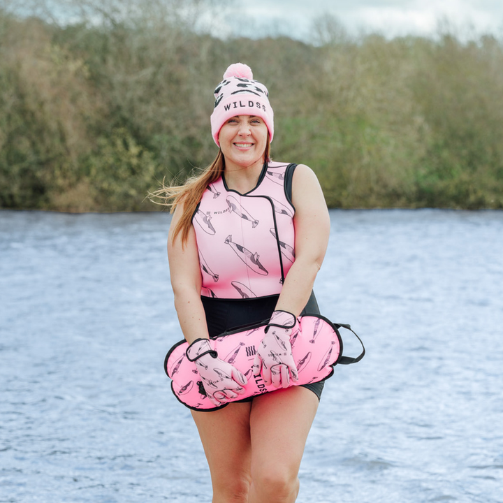Person wearing pink hat, tow float and neoprene swimming gear by water.