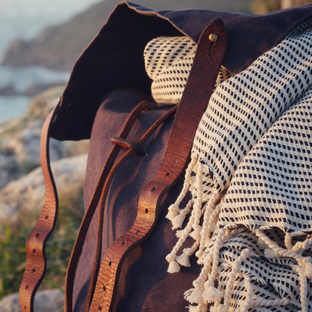 Lightweight cotton Logan hammam towel in a brown leather bag with a coastal scene in the background