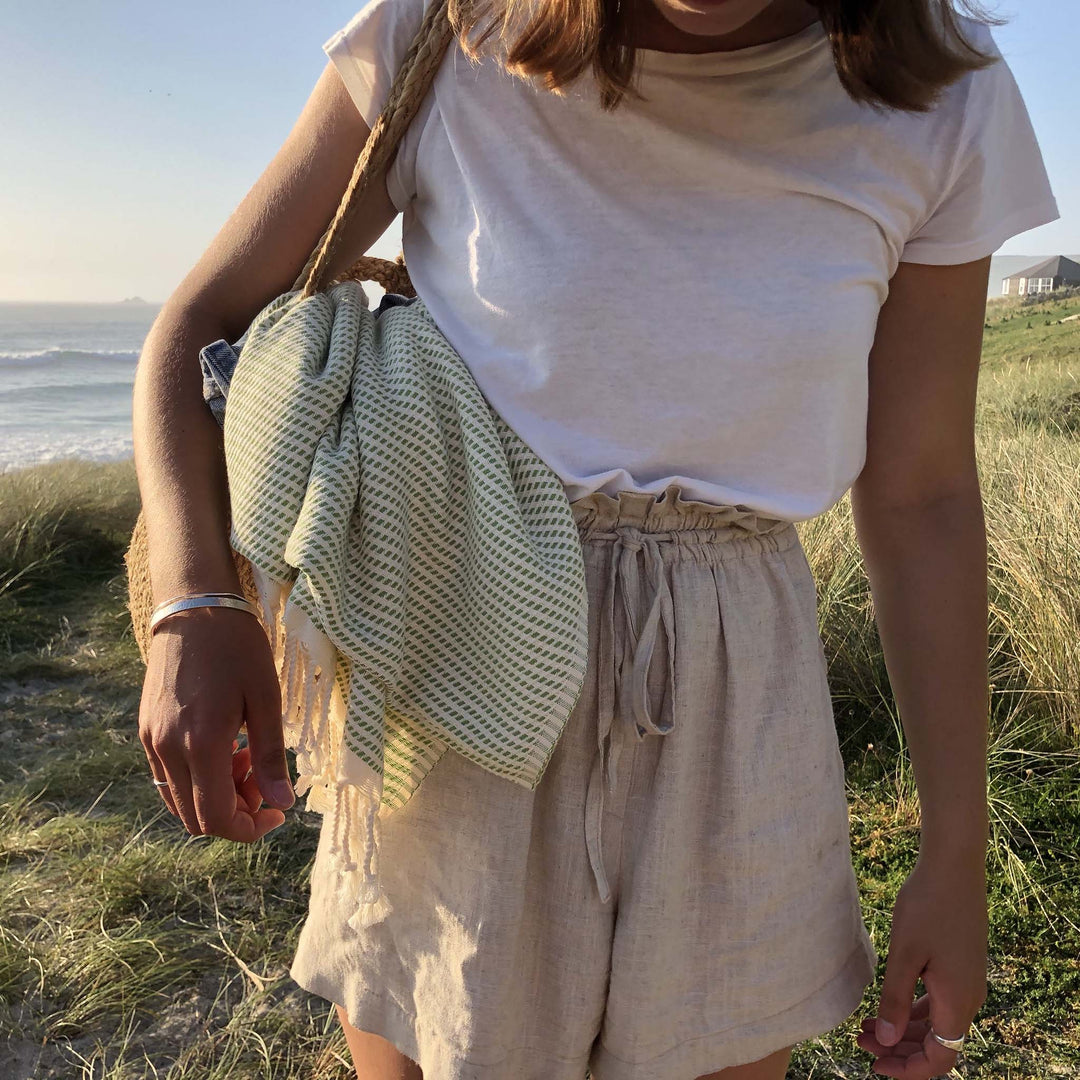 Woman standing in the dunes with a beach bag containing an olive and white stripey cotton hammam towel over her shoulder 