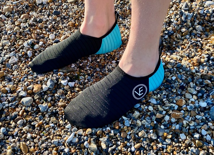 Person on a pebble beach wearing a pair of recyclable adult water shoes with black and aqua upper and black sole, featuring a distinctive VE logo.