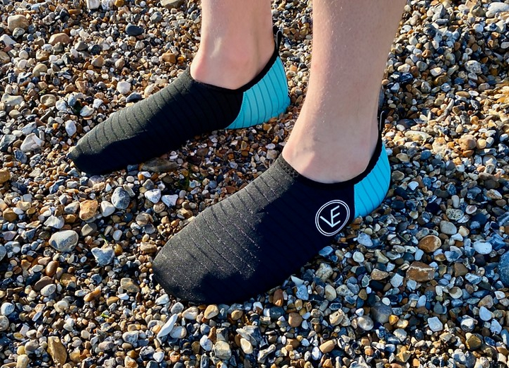 Person on a pebble beach wearing a pair of recyclable adult water shoes with black and aqua upper and black sole, featuring a distinctive VE logo.