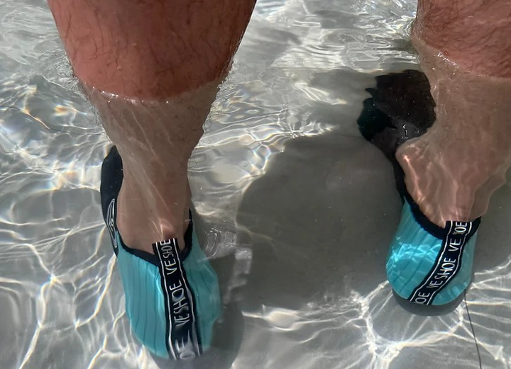 Person wearing a pair of adult water shoes with black and aqua upper and black sole, standing in the water