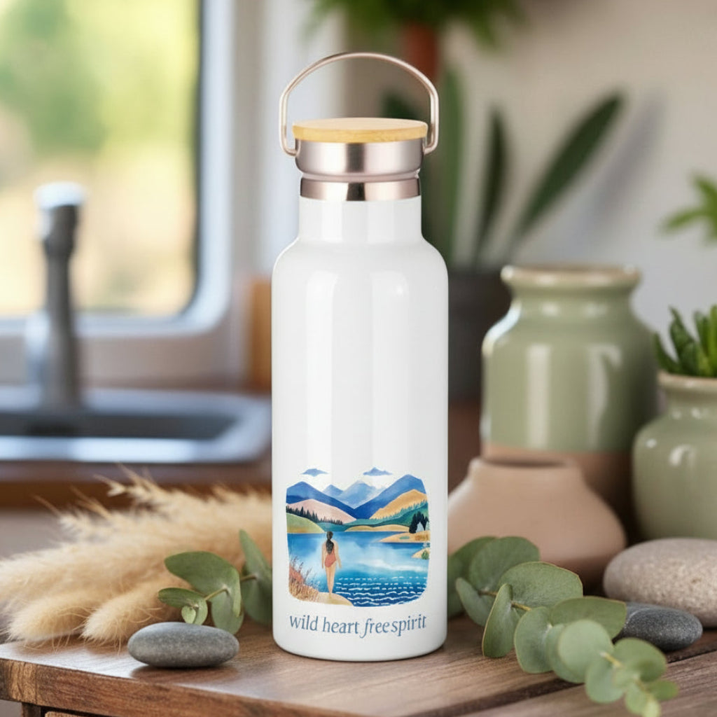 White water bottle with a scenic design and 'wild heart, free spirit' text on a wooden surface with a cup and plants in the background.