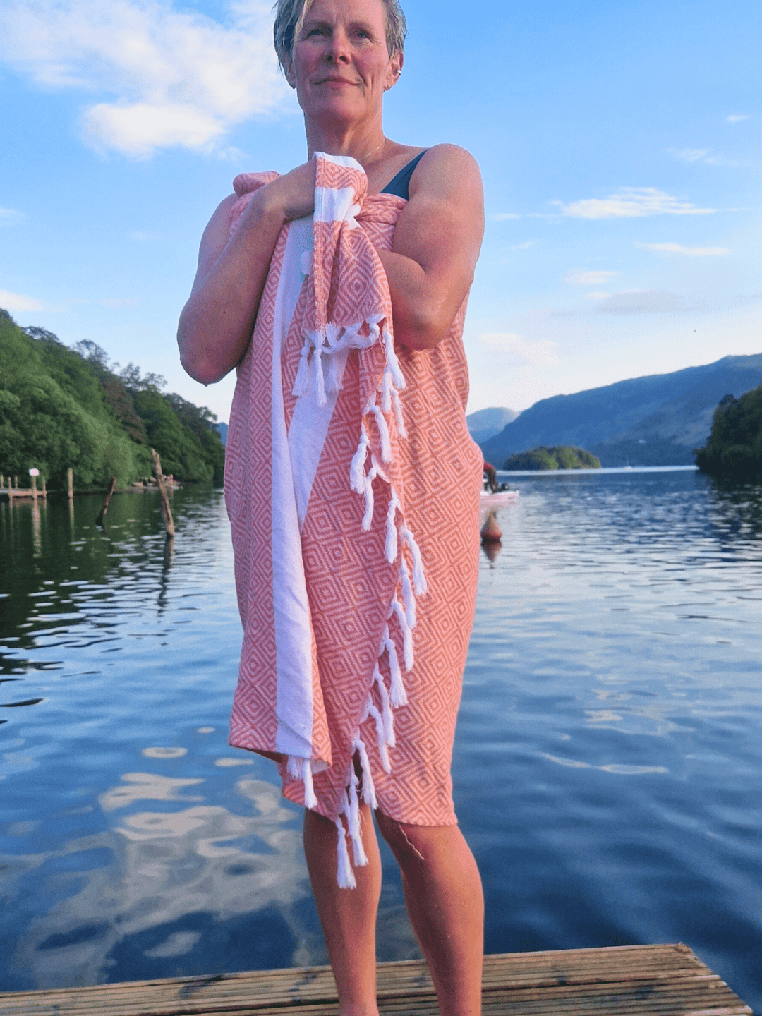 Lifestyle image of an open water swimmer stood on a wooden platform by a lake wrapped up in a Wild Moose hammam towel