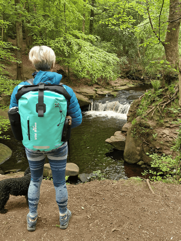 lady with a small black dog wearing the mint green coloured waterproof bag . She is in a  woodland area looking down at a river with a small waterfall