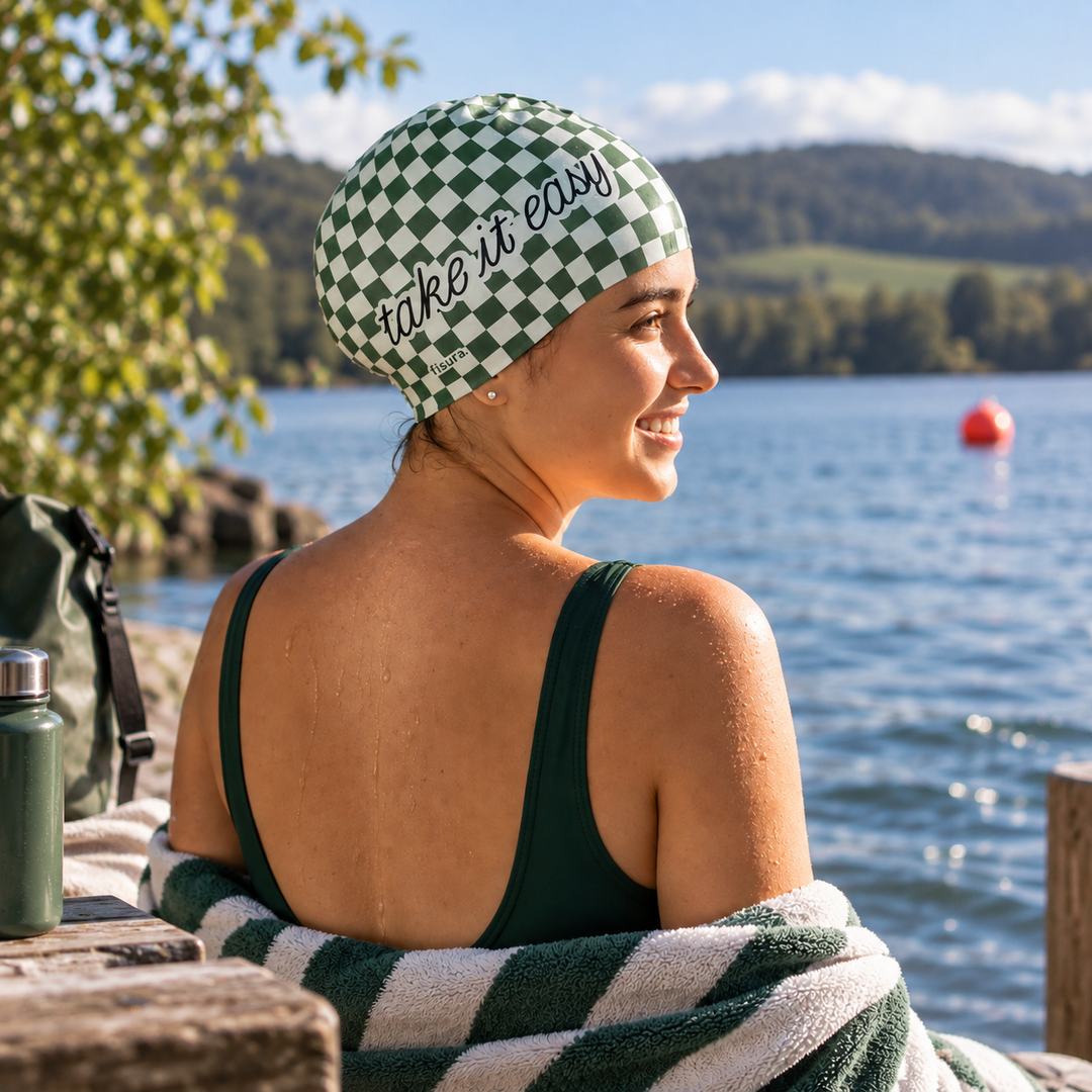A woman sitting by a lake wearing a green and white checkered swim cap with the words 'take it easy' in black script. She is wrapped in a matching striped towel on a wooden dock in the foreground.