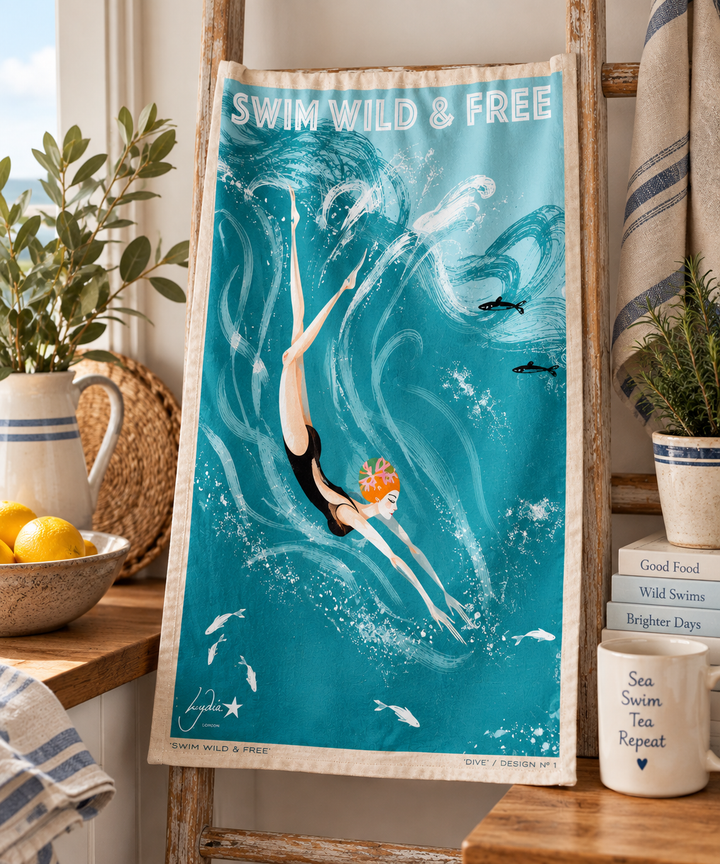 A vintage-style illustrated towel hanging on a wooden ladder in a bright kitchen setting. The towel features a teal and turquoise ocean scene with a diver, fish, and the text &
