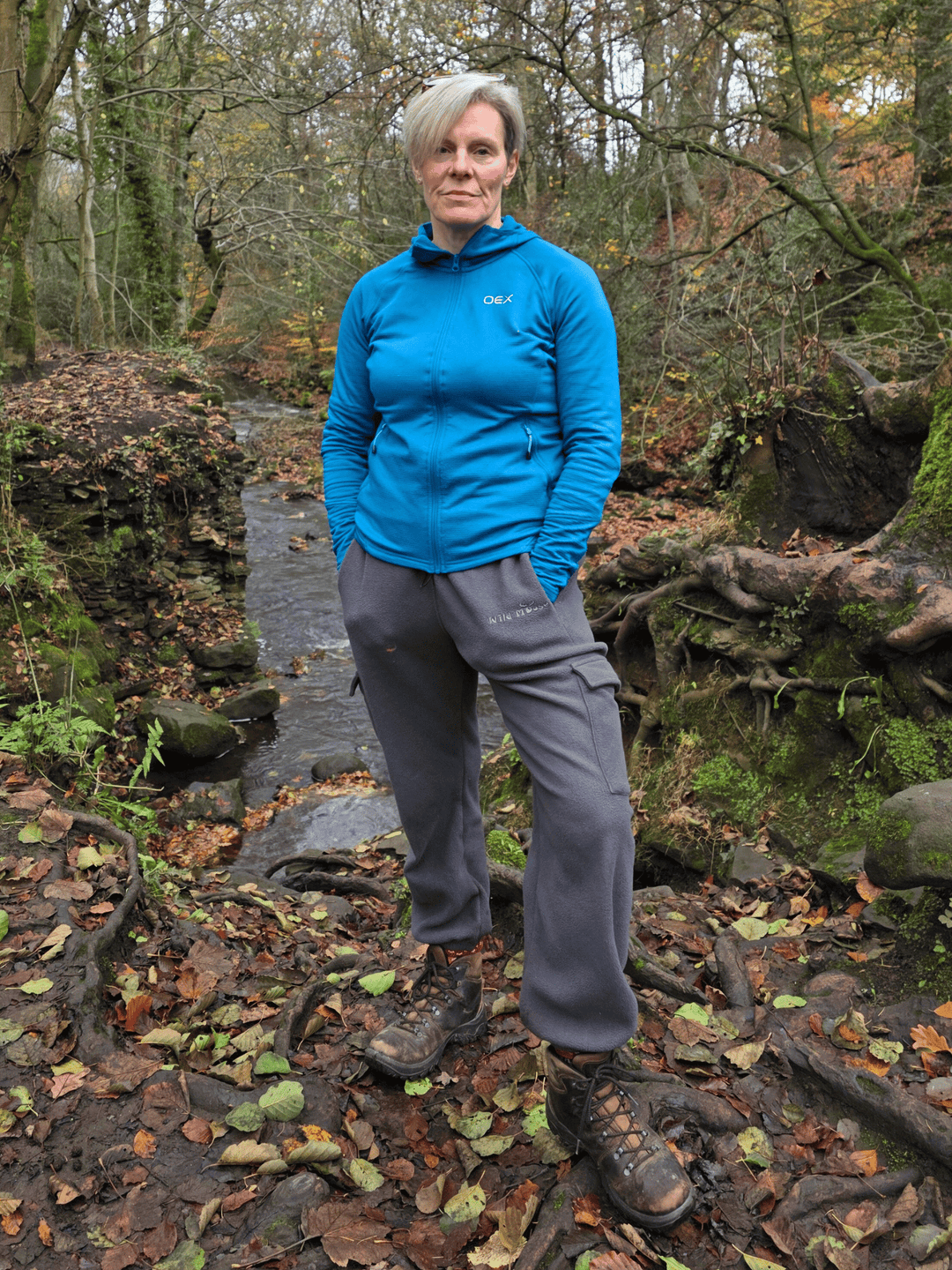 Person wearing  grey fleece jogging pants standing in a forest with a stream.