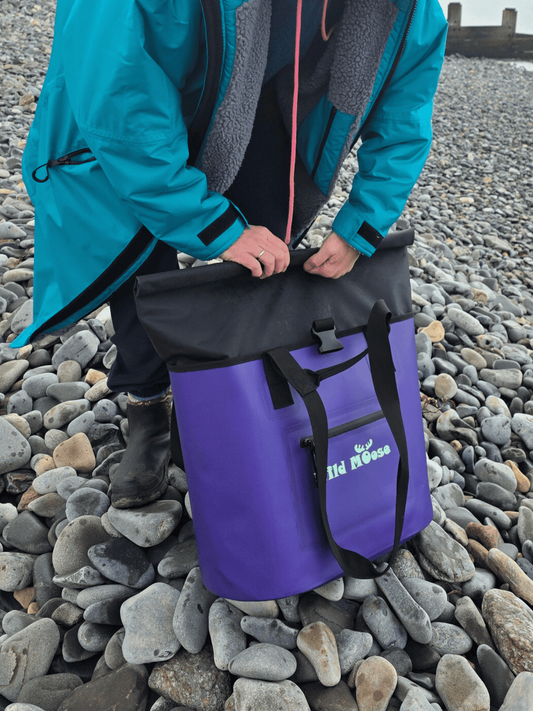 Person unfolding the black top of a purple tote bag on a pebbly beach