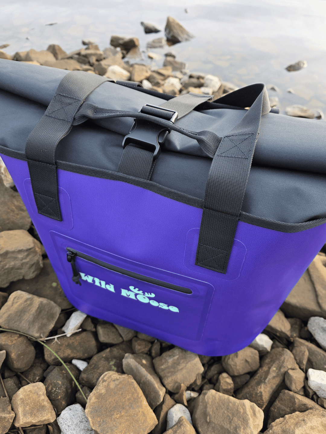 Purple tote bag with black roll top on rocks near water