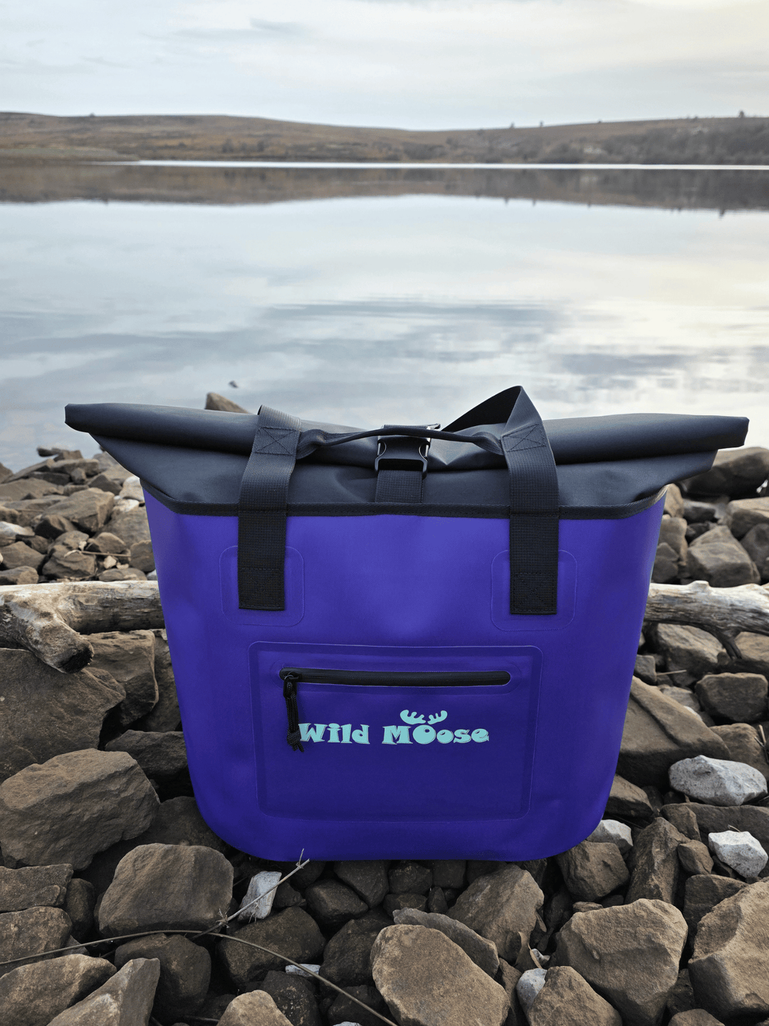 Purple tote bag with black straps on a rocky shore with water in the background