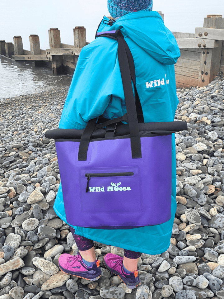 Person wearing a green/blue changing robe and carrying a purple tote bag with &