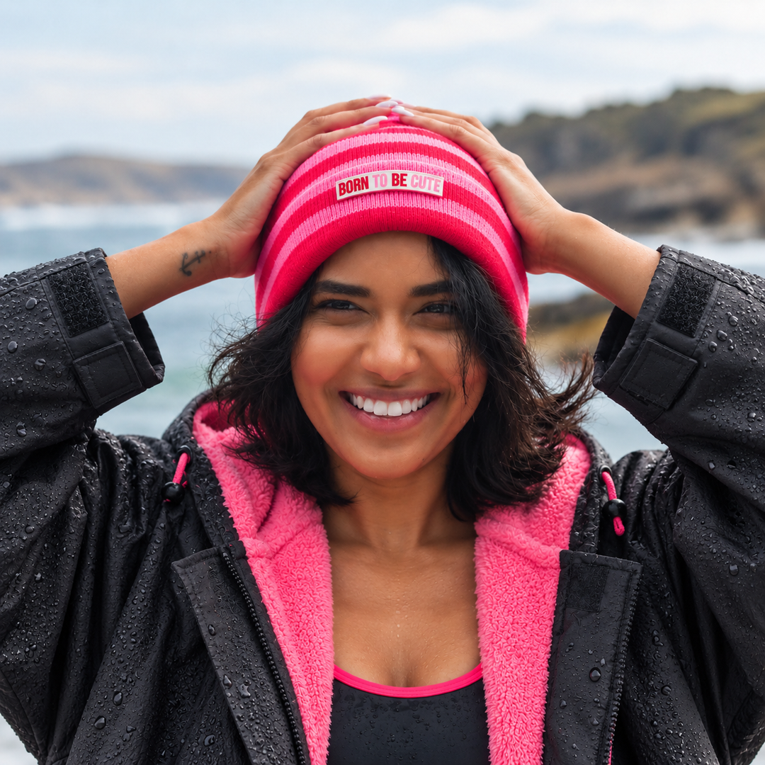 A smiling woman outdoors wearing a bright pink striped knit beanie with a small white rectangular logo that reads 'Born To Be Cute'. 