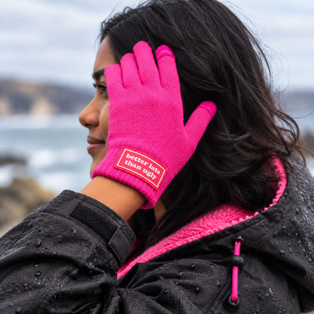 A woman outdoors wearing a bright pink knit glove with a red label that reads 'better late than ugly'. She is also wearing a black open water swimmer changing robe with a bright pink lining.