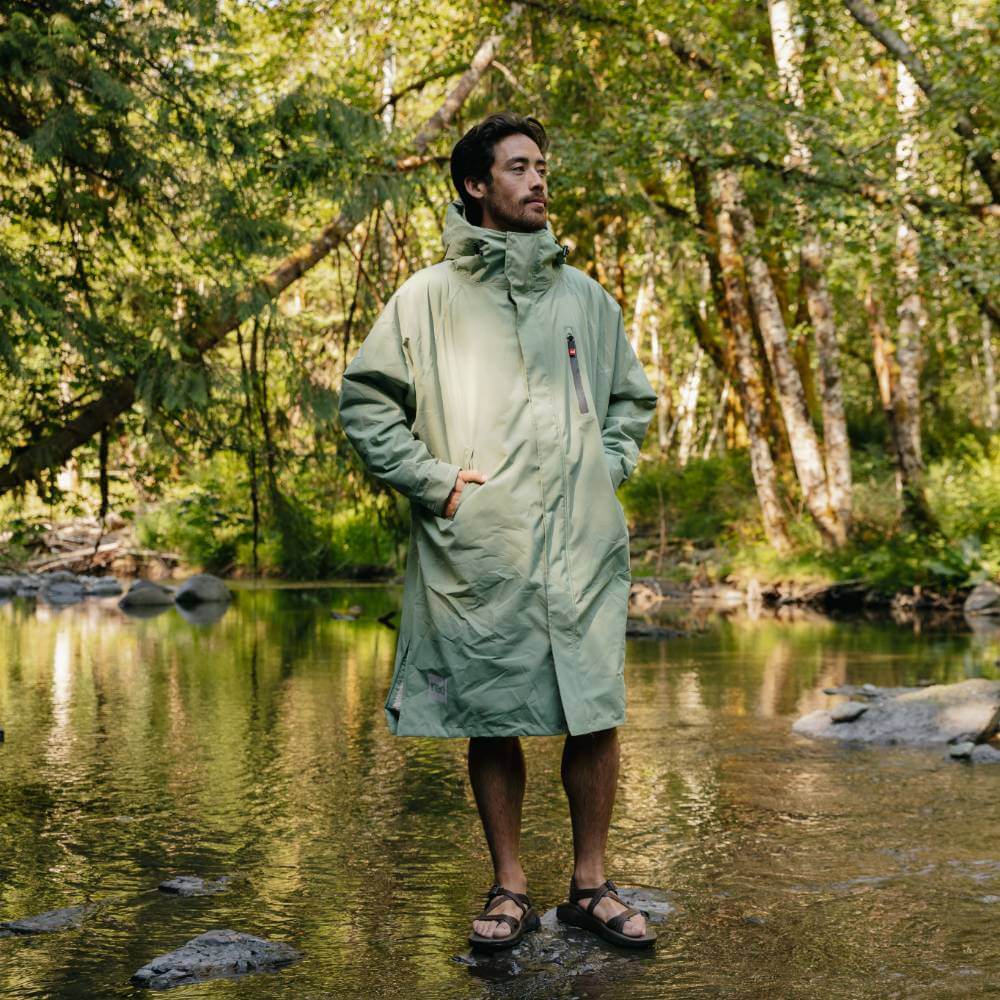 Male swimmer wearing a Red Alter Evo long sleeve waterproof changing robe in Sage Green standing on a stepping stone in the middle of a river.