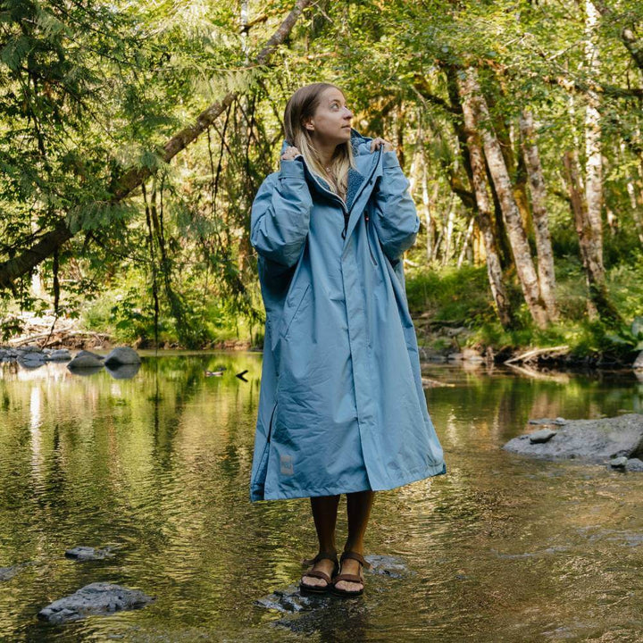 Woman standing on a stepping stone in a river wearing a Red Alter Evo long sleeve waterproof changing robe in Alpine Blue.