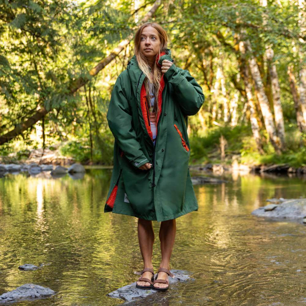 Female swimmer wearing a Red Alter Evo long sleeve waterproof changing robe in Forest Green stood on a stepping stone in the middle of a river.