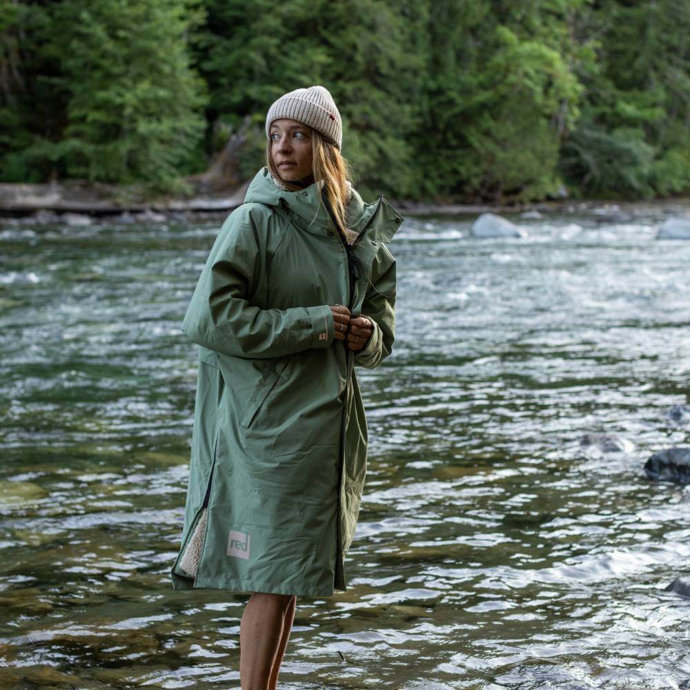 Female swimmer standing on the edge of a flowing river wearing a Red Alter Evo long sleeve waterproof changing robe in Sage Green.