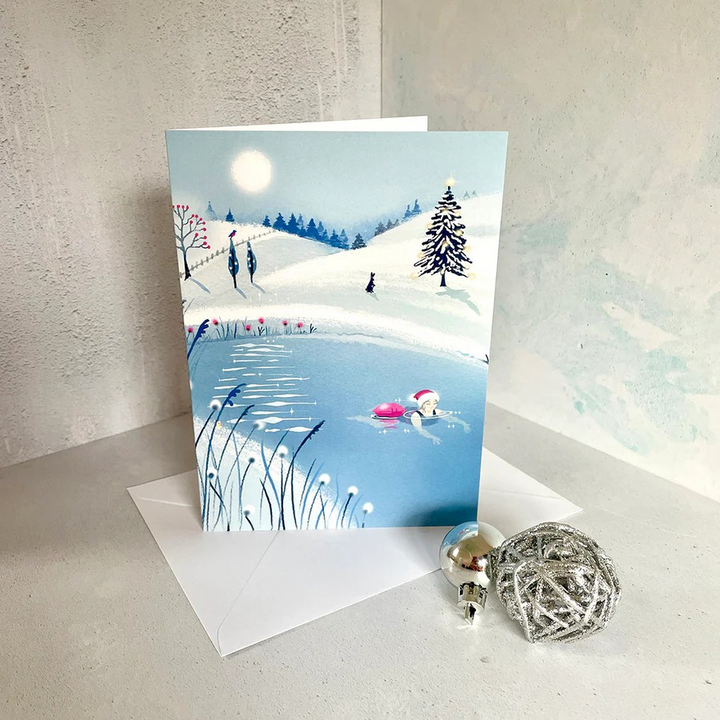 Tis The Season No1 Christmas card by Andrea Hall showing a swimmer in a Santa hat gliding through a frosty pond surrounded by snowy hills and a Christmas tree.