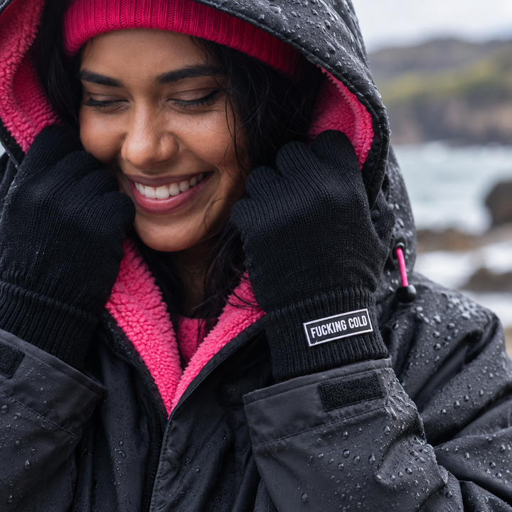 A woman wearing a black changing robe with a bright pink fleece lining and black gloves. The gloves have a small  rectangular patch on the cuff that reads &