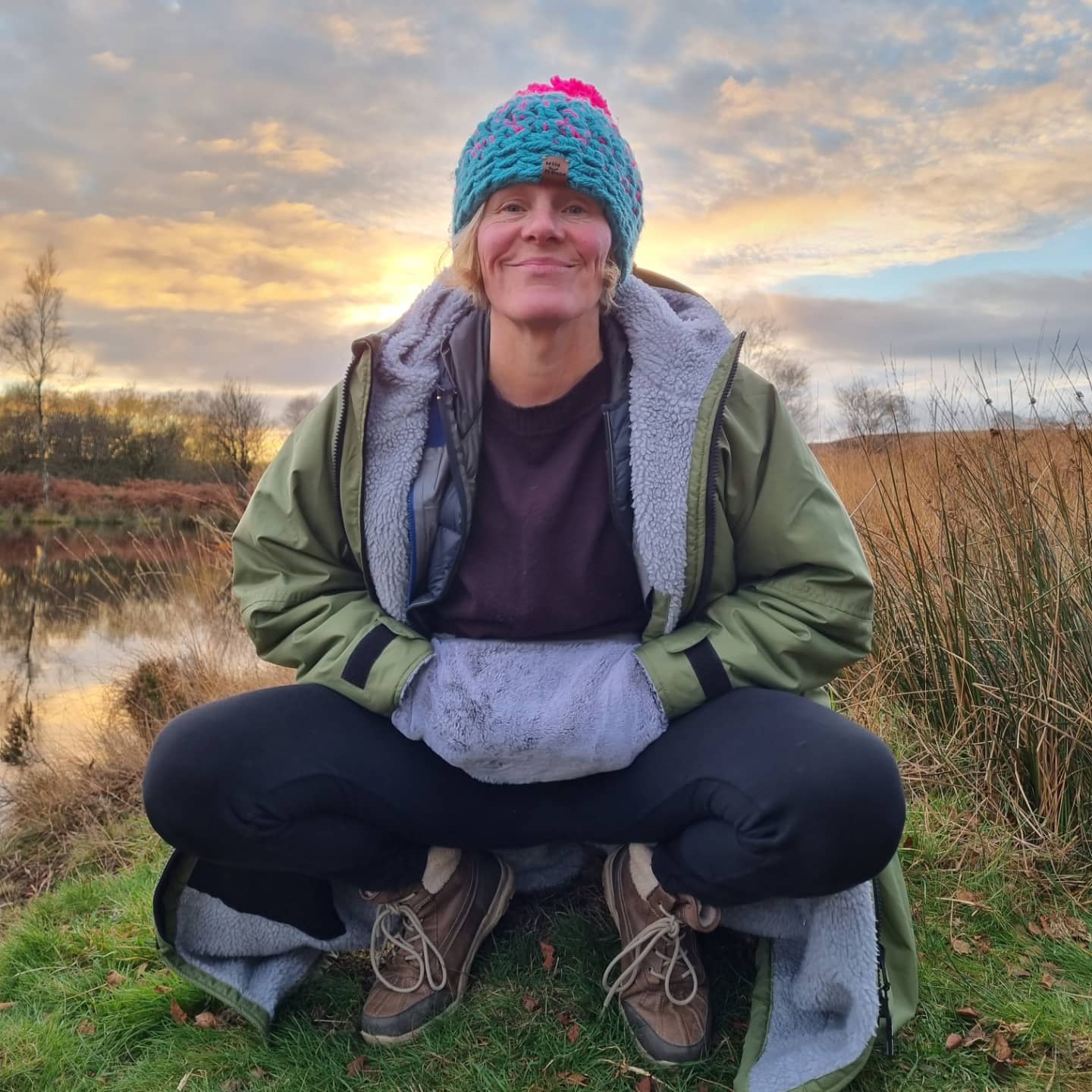 a lady is squatting by a lake in front of a sun  set sky. She is wearing a grey faux fur hottum, a khaki changing robe with a grey lining and a bright colour bobble hat with a big pompom