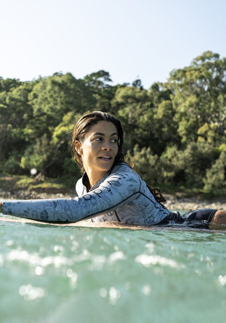 Lifestyle image of a woman on a surf board in the sea wearing the Vivida Linda Reversible Wetsuit 
